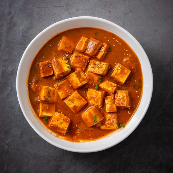A white bowl filled with paneer cubes in a rich orange Indian curry sauce, garnished with seeds.