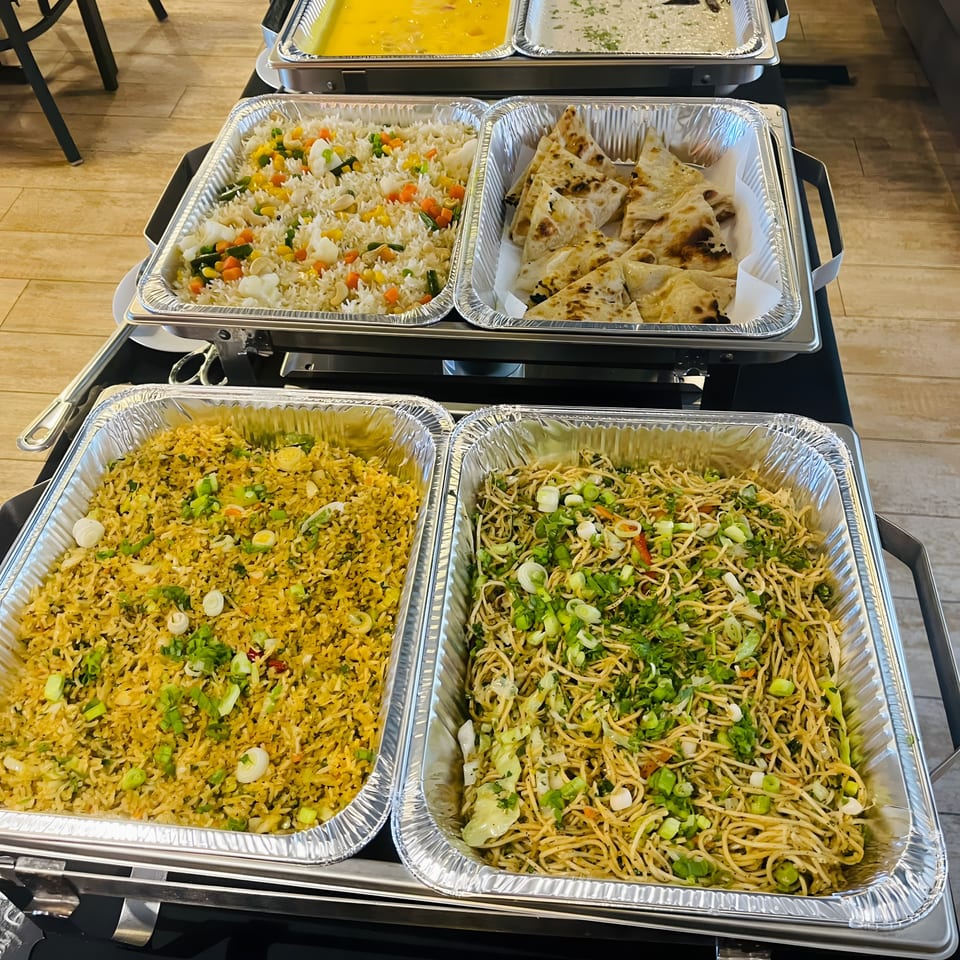 A buffet spread of Indian dishes, including rice, naan, noodles, and curries in foil trays.