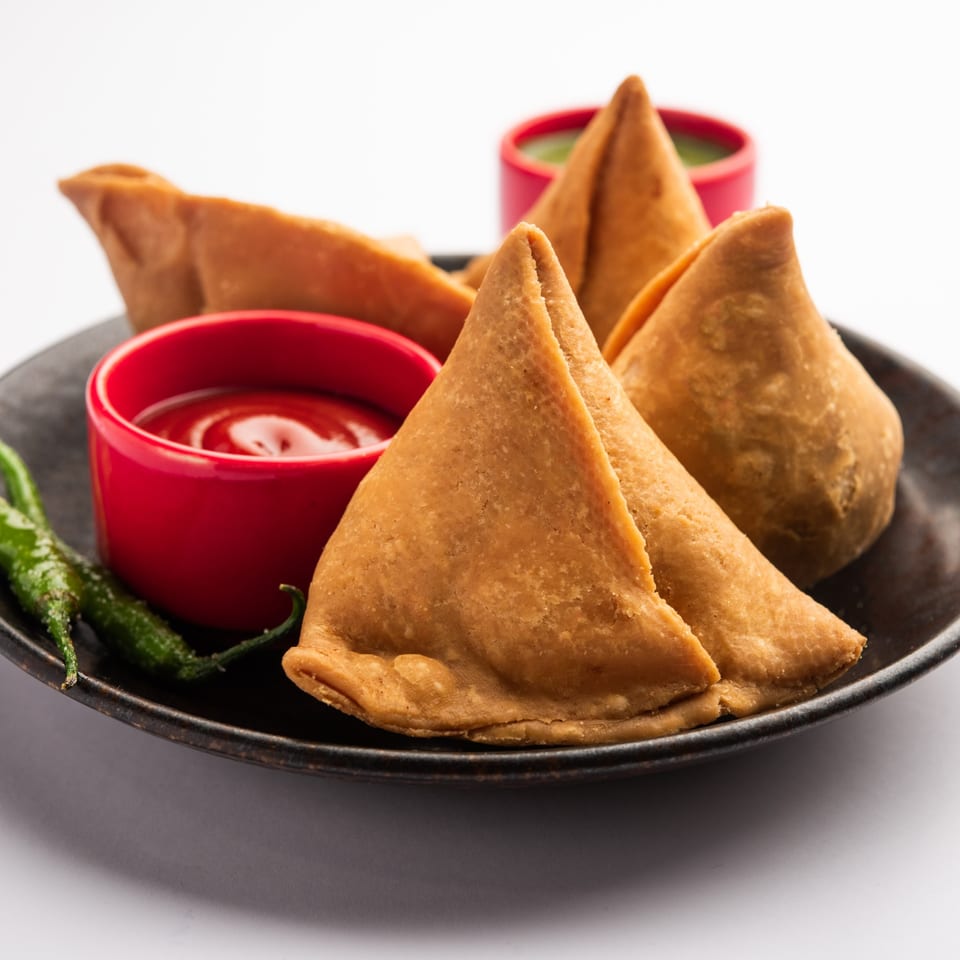 Golden-brown Indian samosas with red ketchup and green chutney in small red bowls, garnished with green chilies.
