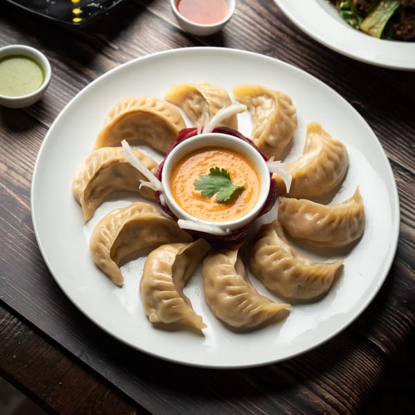 A white plate of steamed momos with a central bowl of orange dipping sauce, garnished with cilantro.