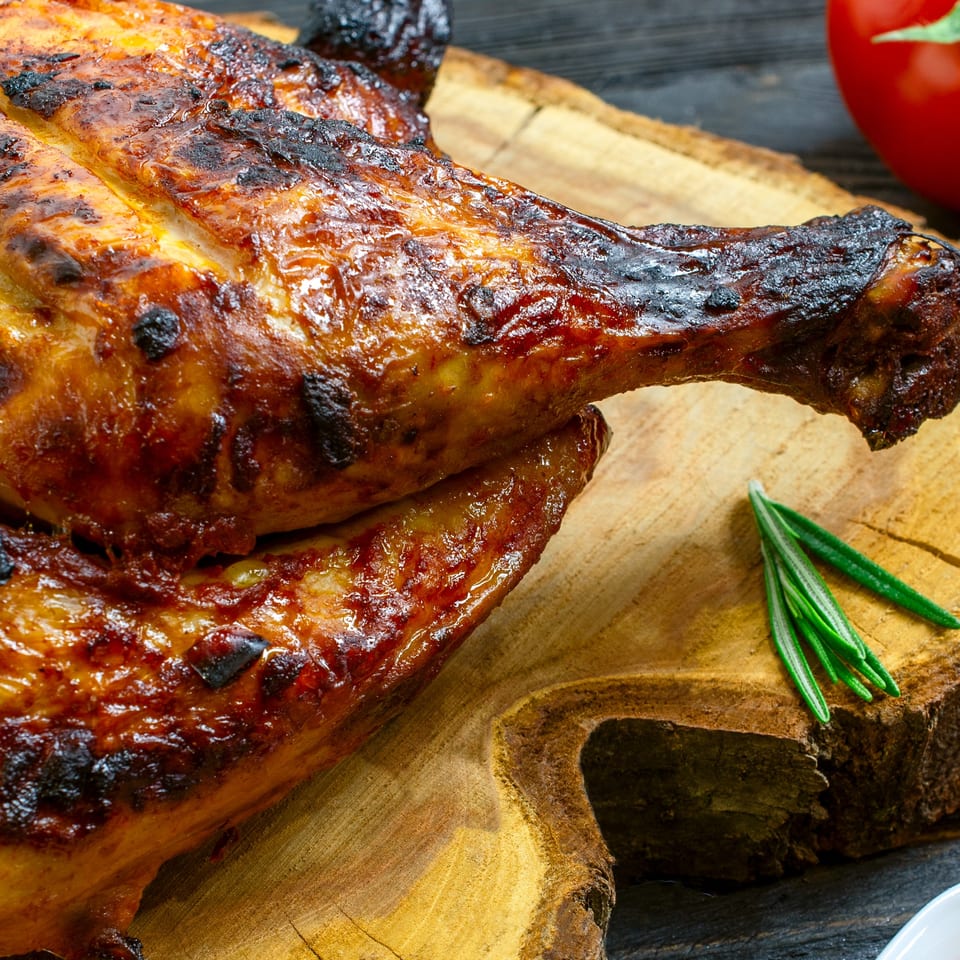 Close-up of a whole roasted chicken with crispy skin, rosemary, and a tomato on a wooden board.